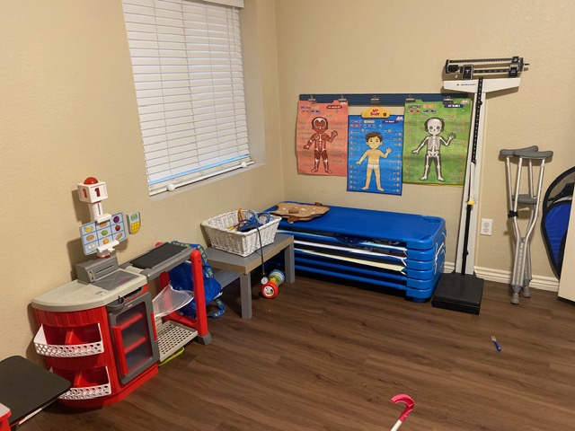 Playroom with a toy kitchen set, stacked blue mats, anatomy posters on the wall, a medical scale, and various toys on a wood floor.