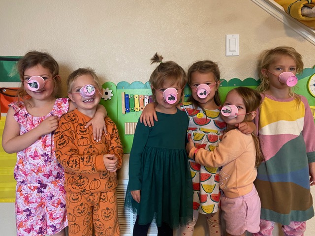 Six children wearing pig nose masks stand in a classroom, some with arms around each other, smiling at the camera.