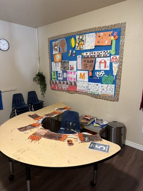 A classroom art area with a U-shaped table, blue chairs, children's artwork on the wall, a clock, books, and a hanging plant.