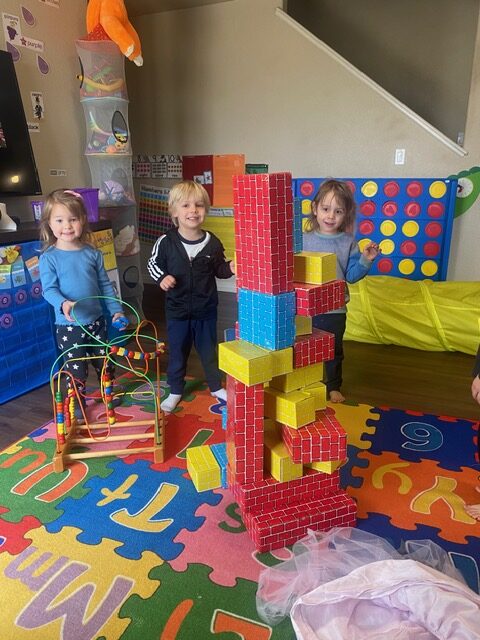 Three young children stand in a playroom with colorful foam mats, toys, and a tall structure built from large toy blocks in the center.