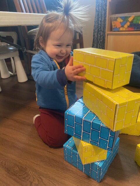Toddler in blue and red clothes stacking large yellow and blue toy blocks on a wooden floor, smiling and focused.