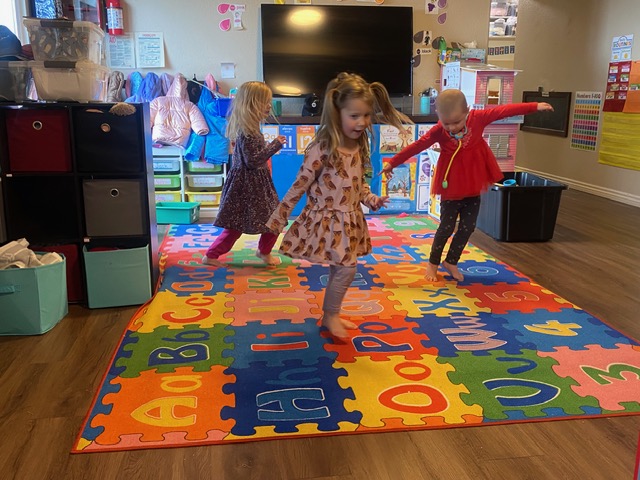 Three young children play and move energetically on a colorful alphabet mat in a classroom with storage bins and educational posters.